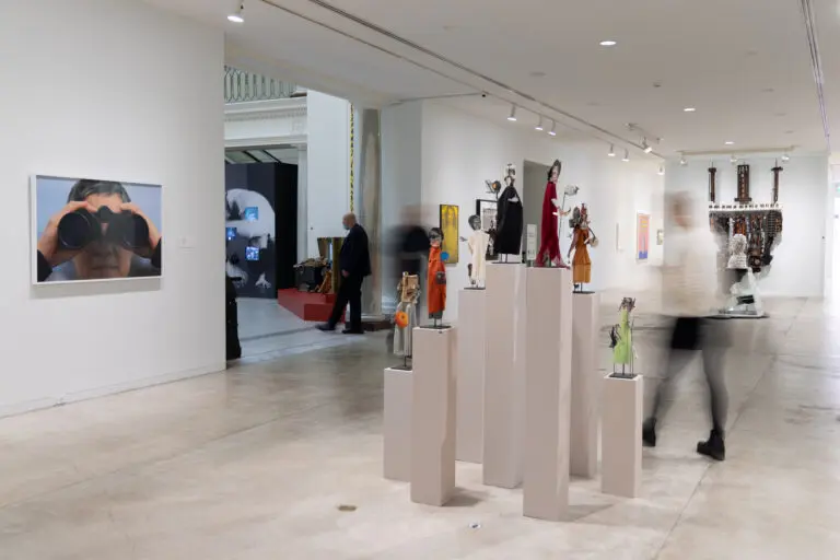 A photograph of a blurred person walking through a white-walled gallery with different artwork on view including photographs, collage sculptures on plinths and a deconstructed piano sculpture in the background.