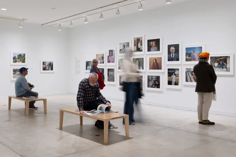 A photograph of a white-walled Gallery with white framed colour photographs on the wall. People stand and look at the photos. Others sit and look or read materials on two benches in the middle of the room.
