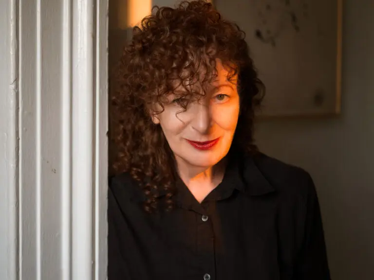 A photo portrait of artist Nan Goldin. She stands next to a wall, and the glow of sunlight appears across half her face. She has curly, red hair.