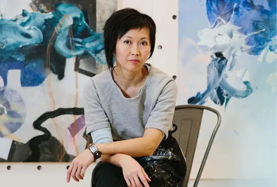 A photograph of artist Vanessa Lam sitting on a chair in her studio with large scale, abstract paintings in blue hues behind her.