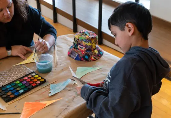 A photograph of a young boy in a sweatshirt using scissors to cut out a shape in paper. He stands at a making station table and works with a woman across from him.