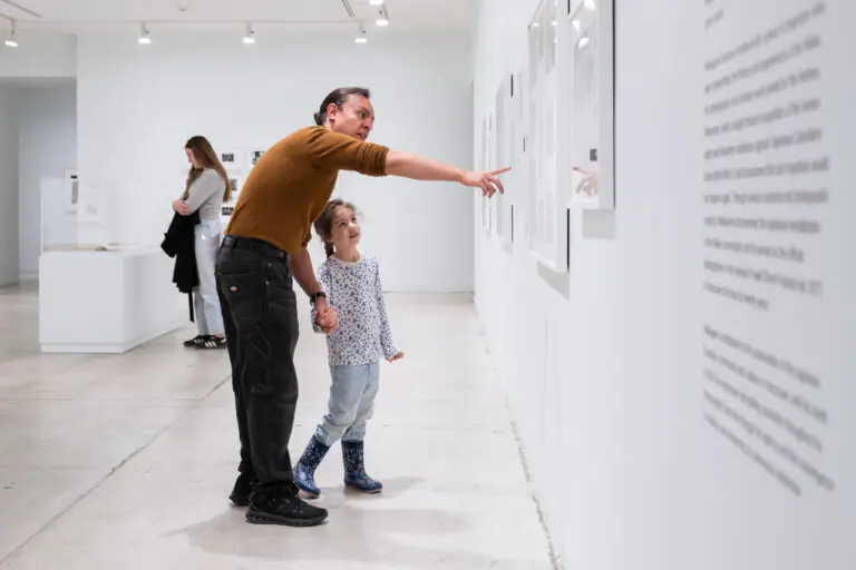 A photo of a father and daughter holding hands and looking at art in a white-walled gallery. The father is pointing at an artwork on the wall. A woman is standing in the background.