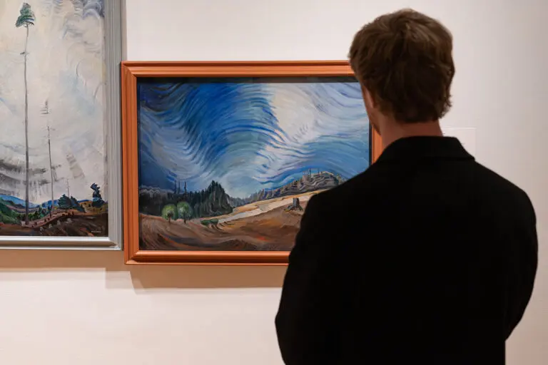 A photograph of a man from behind, from the torso up, looking at a painting by Emily Carr of a horizontal landscape.