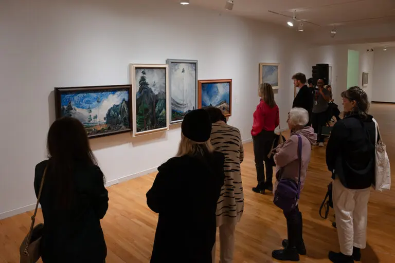 A photograph of a group of people standing in the Gallery looking at a wall of Emily Carr paintings.