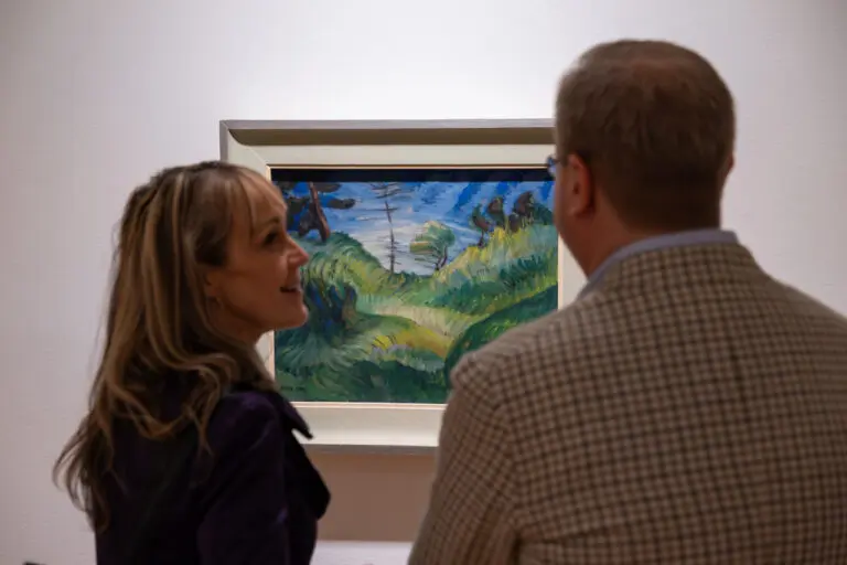 A photograph of two people, a man and a woman, from the chest up, looking at a painting of a forest landscape painted by Emily Carr. The people's backs are shown to viewers, but the woman turns to the man to say something.