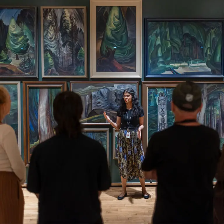 A group of men and women visit the Gallery and enjoy a guided tour. They are standing with their backs to us, looking at a dark green wall with Emily Carr paintings of the forest on it. A young woman tour guide stands in front of the paintings gesturing and looks back at the group.