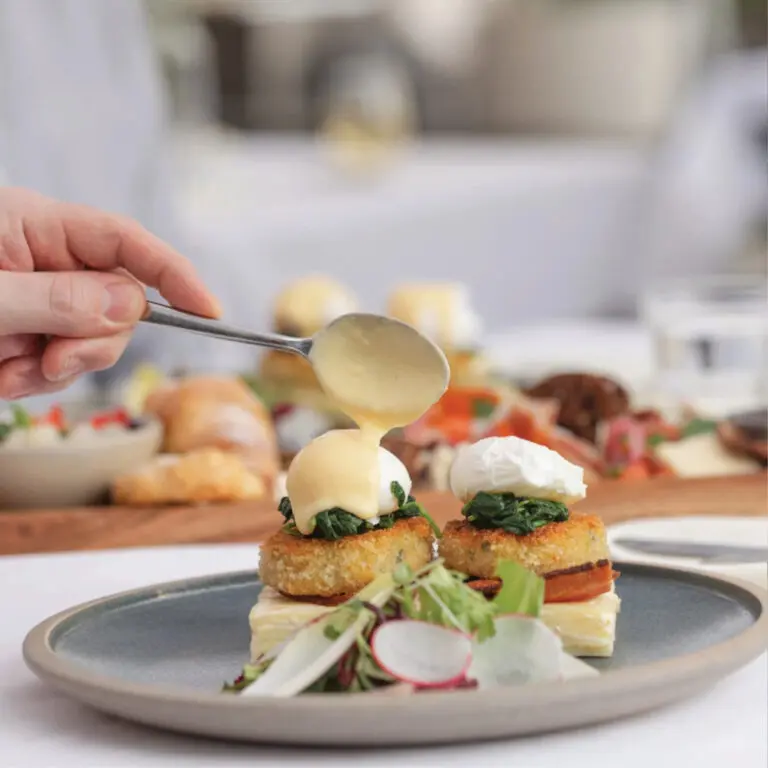 A square image of a hand pouring hollandaise sauce onto eggs benedict on a plate.