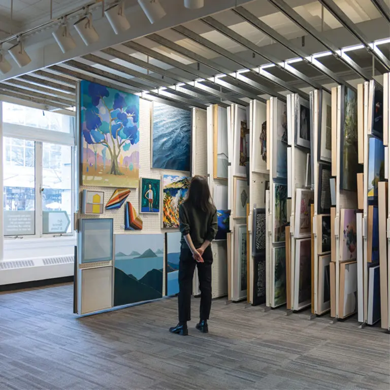 A square photograph of a woman from behind standing and looking at a pull out of artworks on a rack. There are rows of closed artworks racks in the room.