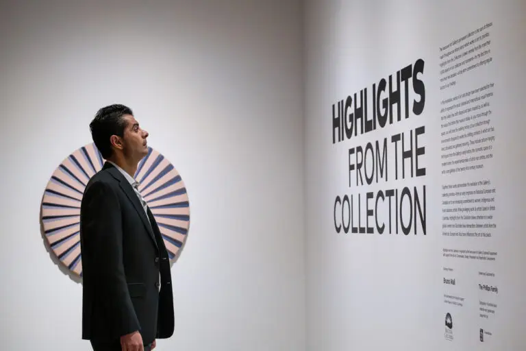 A photograph of a man from the side standing in front of the title wall in an exhibition space. The exhibition title appears on the grey wall in black: Highlights from the Collection. Behind the man is a circular sculpture on the wall in pink and purple.