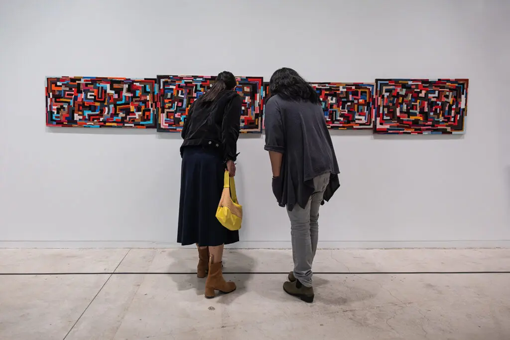 A photograph of two people from behind leaning in to take a closer look at a multicoloured, rectangular work of textile art on the wall in a gallery.