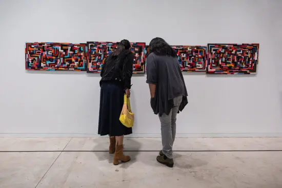 A photograph of two people from behind leaning in to take a closer look at a multicoloured, rectangular work of textile art on the wall in a gallery.