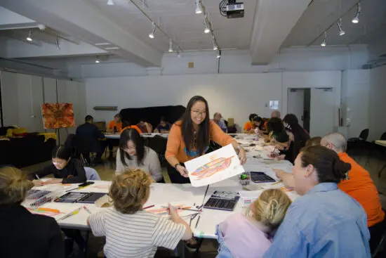 A photograph of an Eagle Feather Design Workshop. There is a room of families sitting at tables arranged in a u shape. An Indigenous woman wearing an orange shirt stands in the centre of the tables and leans over to give an eagle feather drawing to a family.