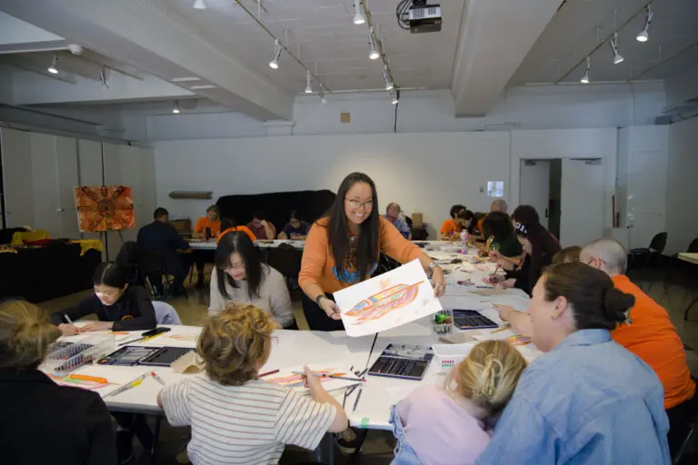 A photograph of an Eagle Feather Design Workshop. There is a room of families sitting at tables arranged in a u shape. An Indigenous woman wearing an orange shirt stands in the centre of the tables and leans over to give an eagle feather drawing to a family.