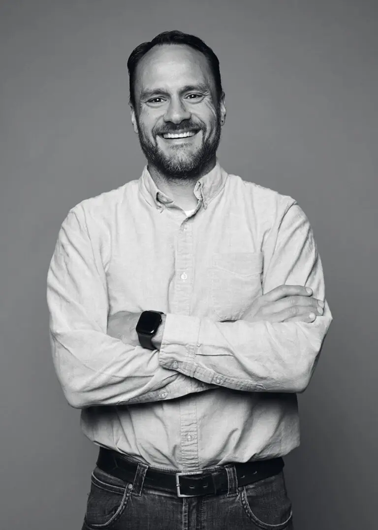 A black and white photograph of a man in a white collared, button up shirt from the waist up. He has a short beard and a large, welcoming smile on his face. His arms are crossed in front of him.