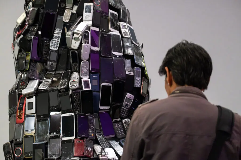 A photograph of a person from behind from the shoulders up. They are looking at a sculpture made up of old cell phone waste.