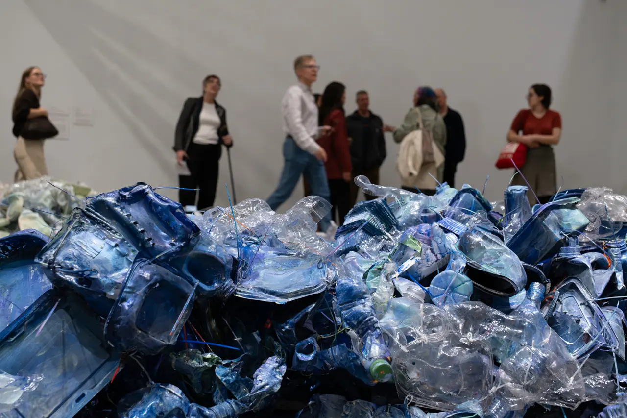 A photograph detailing an artwork by Liz Larner of plastic waste from the ocean, primarily water bottles, painted blue. Behind the wall of plastic in the foreground of the image, there are people mulling about in the gallery space.