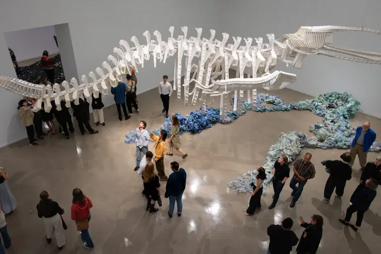 A photograph taken from above of a Gallery room featuring a large whale skeleton made out of white plastic chairs. There are people in the room looking up in awe at the work. On the floor, there is another installation that recalls the ocean made up of painted blue plastic bottles.