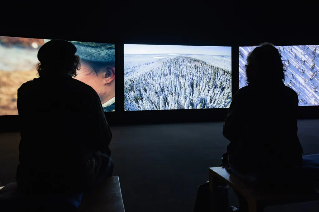 A photograph of two silhouettes of people from behind in a dark room watching a three-channel video projection. In the centre is an image of a mountain covered in snow.