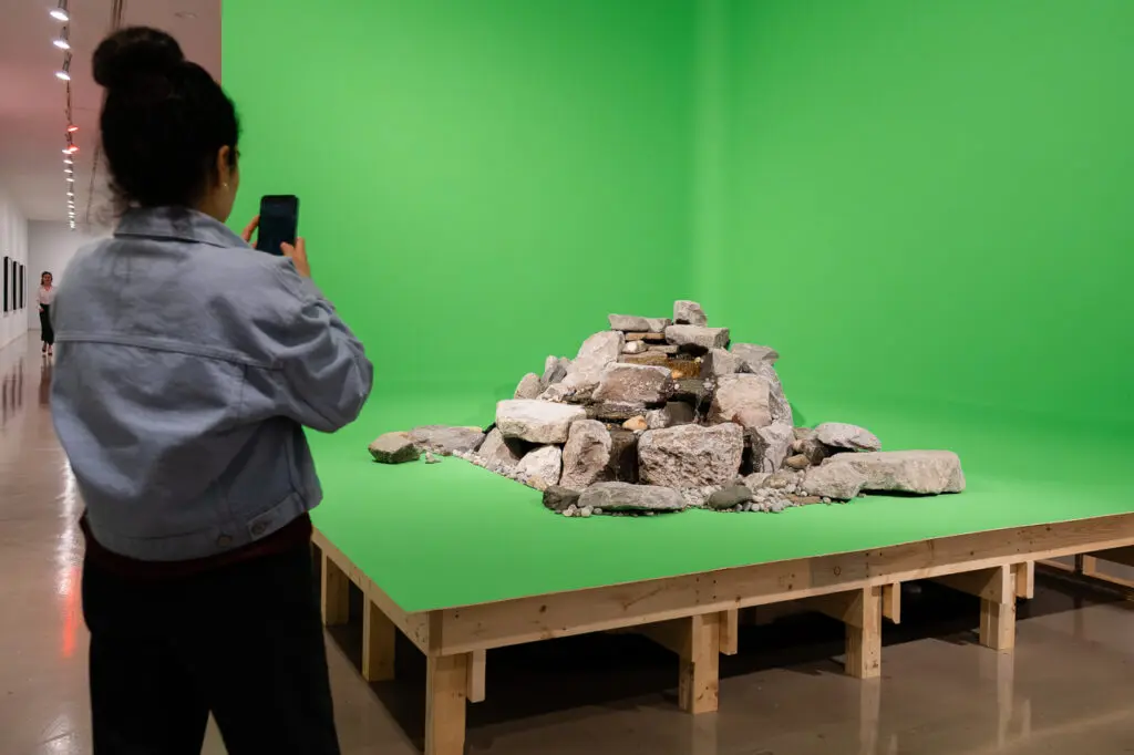 A photograph of a woman from behind who is standing and taking a photograph on her phone of a large set-like structure on wooden legs, with a green background. In the middle there is a pile of rocks.
