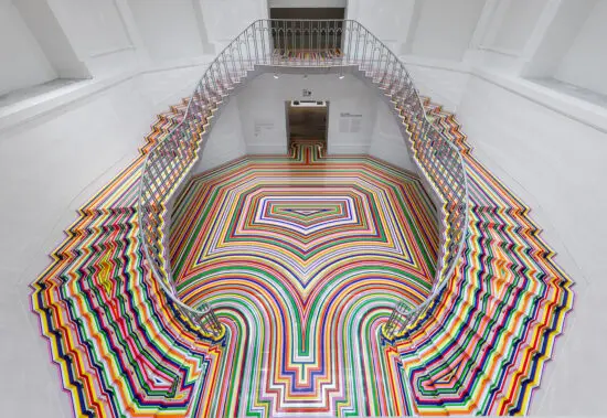 An installation photograph of Jim Lambie's artworks Zobop (Colour-Chrome) and Zobop (Colour-Chrome) Stairs covering the stairs and floor of the Vancouver Art Gallery's neo-classical Rotunda. The floor and stairs are covered in brightly coloured strips of vinyl in purple, pink, green, red, orange and yellow.