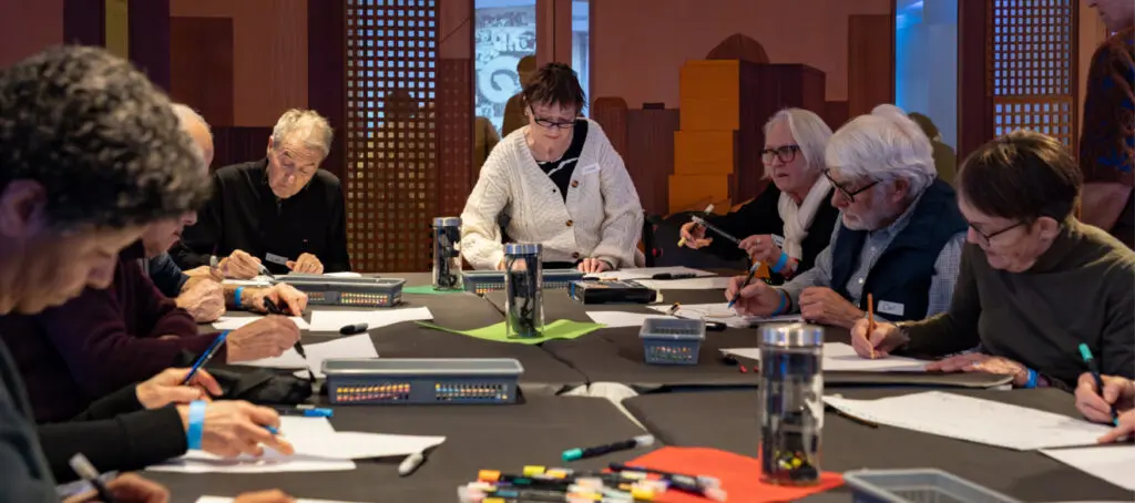 A photograph of a group of seniors, men and women, seated at a table during a workshop. They are drawing.
