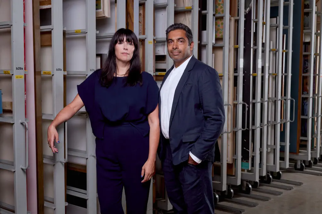 A photograph of a Eva Respini and Sirish Rao standing in the Gallery's vault. A woman stands with her arm leaning against the racks of art. A man stands next to her with his hands in his pockets.