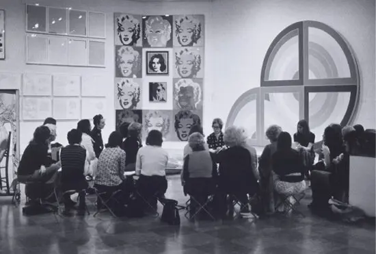 An archival black-and-white photograph of a group of docents sitting around curator Doris Shadbolt in an exhibition space as they get a briefing.