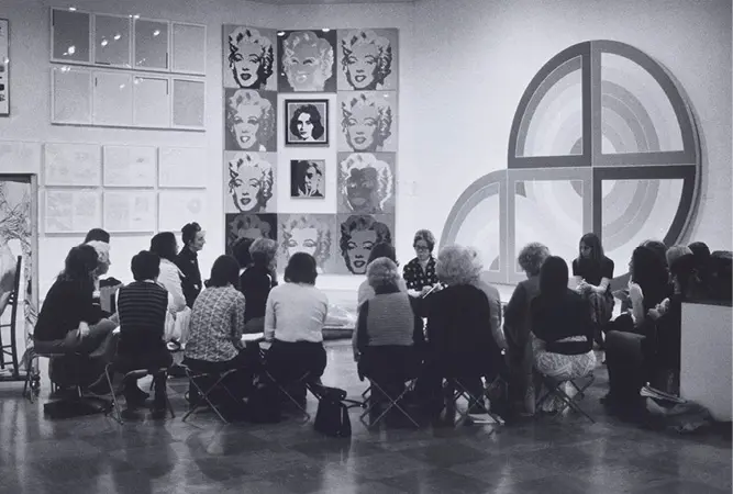 An archival black-and-white photograph of a group of docents sitting around curator Doris Shadbolt in an exhibition space as they get a briefing.