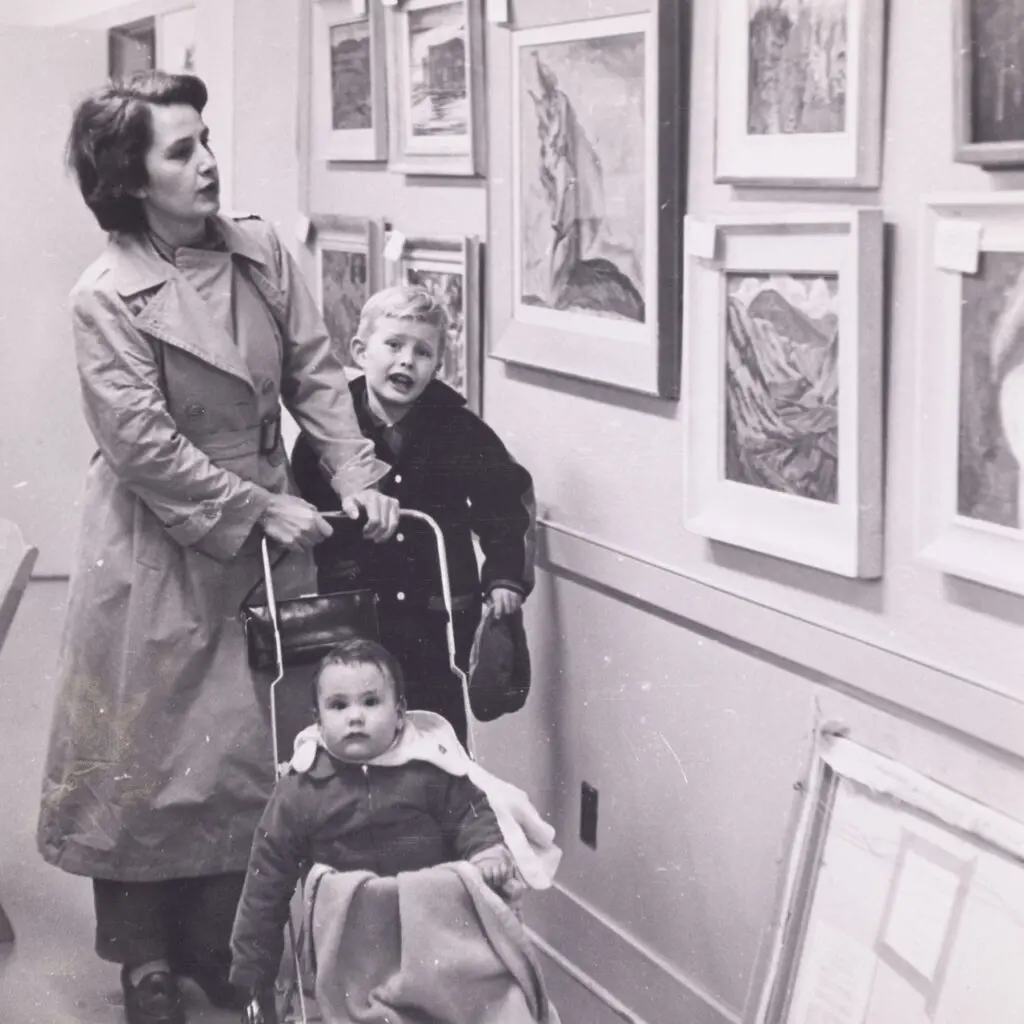 An archival photograph in black and white of a mother with a child and a baby in a pram looking at artworks.