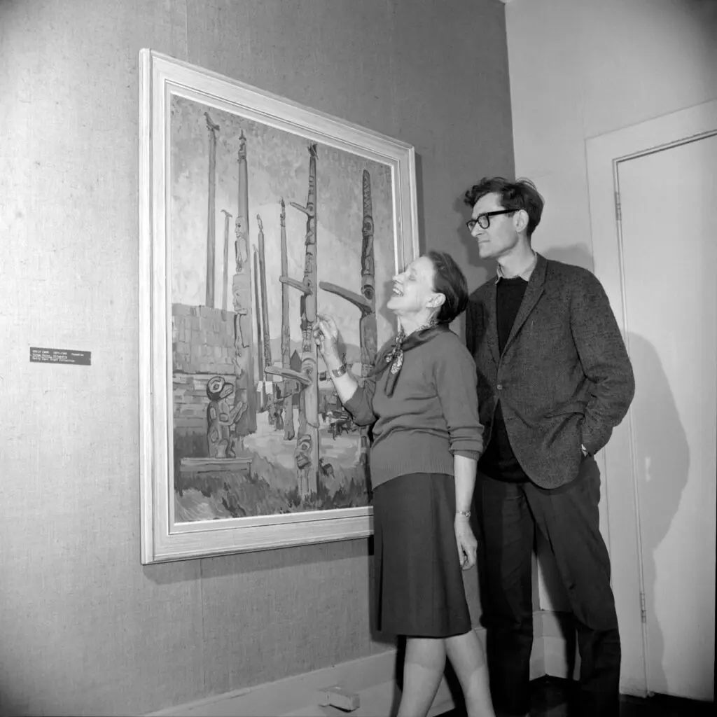 An archival photograph of a man and a woman looking at a painting by Emily Carr of totem poles.