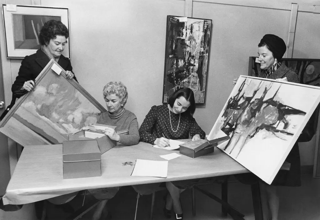 An archival photograph in black and white of 4 women in an office looking at pieces of art.