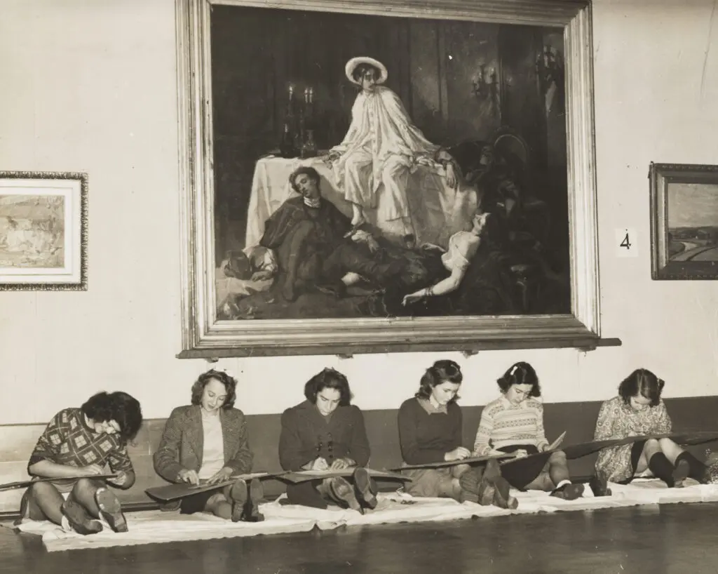 An archival photograph in black and white of 6 students sitting on the floor of the Gallery below a painting as part of a class visit.