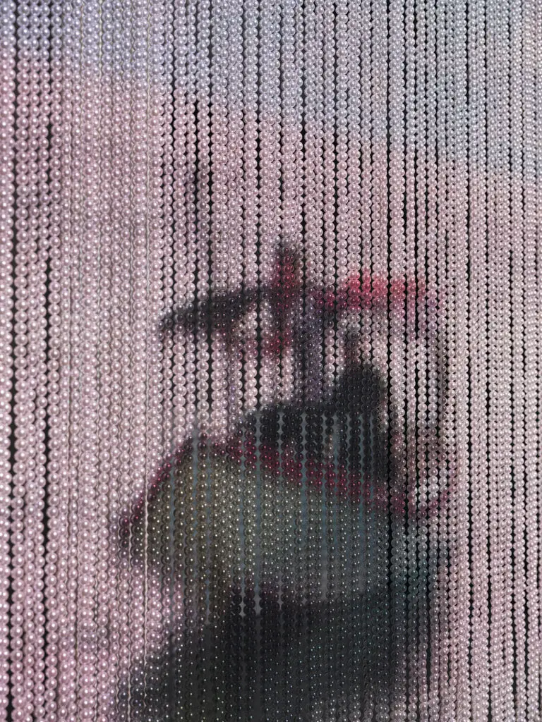 A photograph of a video artwork. An image of a boat with a person it in is projected on a wall of stringed white pearls.