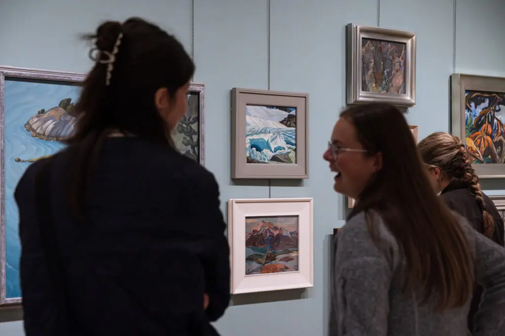 A photograph with two women in the foreground, slightly out of focus. They are looking at each other talking and smiling. Beyond them we see paintings of landscapes hanging on the wall.