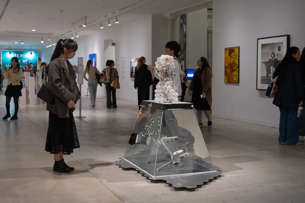 A photograph of an exhibition gallery with lots of people mulling about. At the centre is a woman looking at a sculptural work that appears to be made up of lightbulbs.