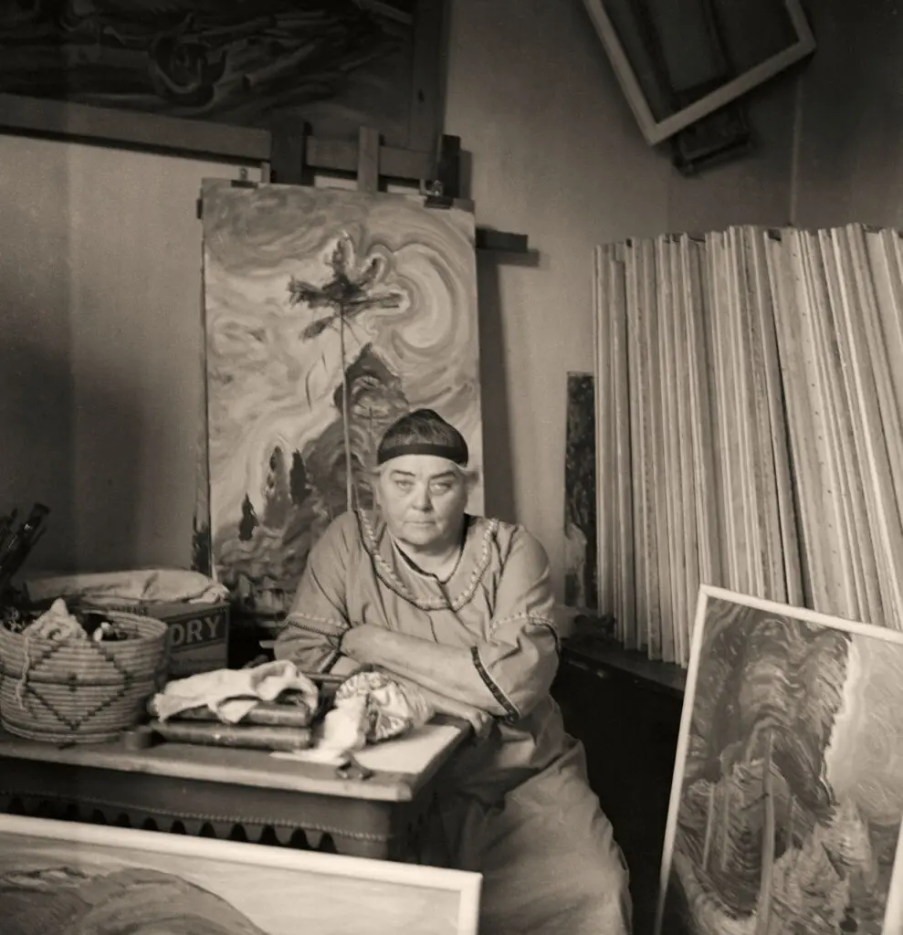 A photograph in black and white of Emily Carr in her studio surrounded by paintings.
