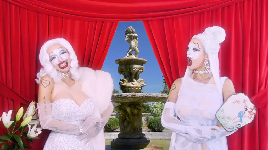 A video still from an art film featuring two women in pink dresses wearing blonde wigs standing in from of a theatrical backdrop of red curtains and a fountain.