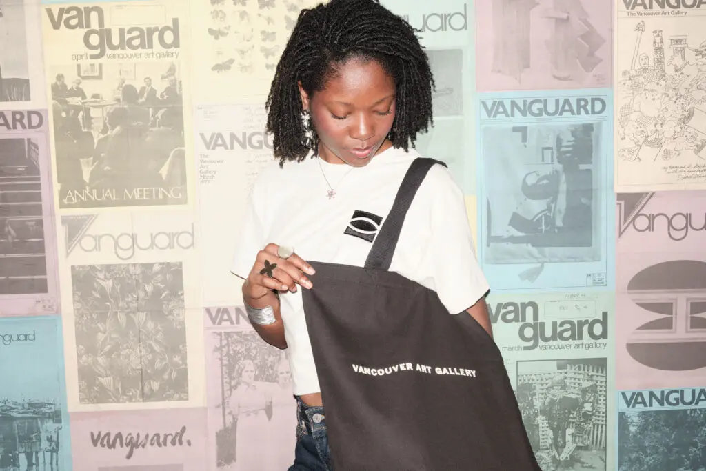 A photograph of a young Black woman wearing a black tote bag that reads Vancouver Art Gallery and an ecru t-shirt with a black eye logo on it. She stands in front of a wall of posters of historical magazine covers.