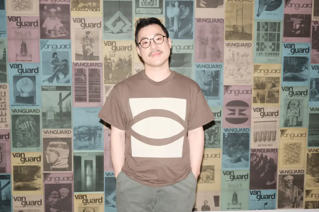 A photograph of an Asian man with short black hair wearing an olive t-shirt with the eye graphic logo on it. He stands in front of a wall of posters of historical magazine covers.
