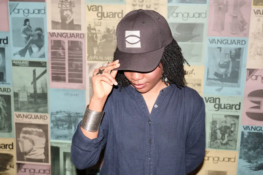 A photograph of a Black woman with wearing a cap with the eye logo on it. She stands in front of a wall of posters of historical magazine covers.