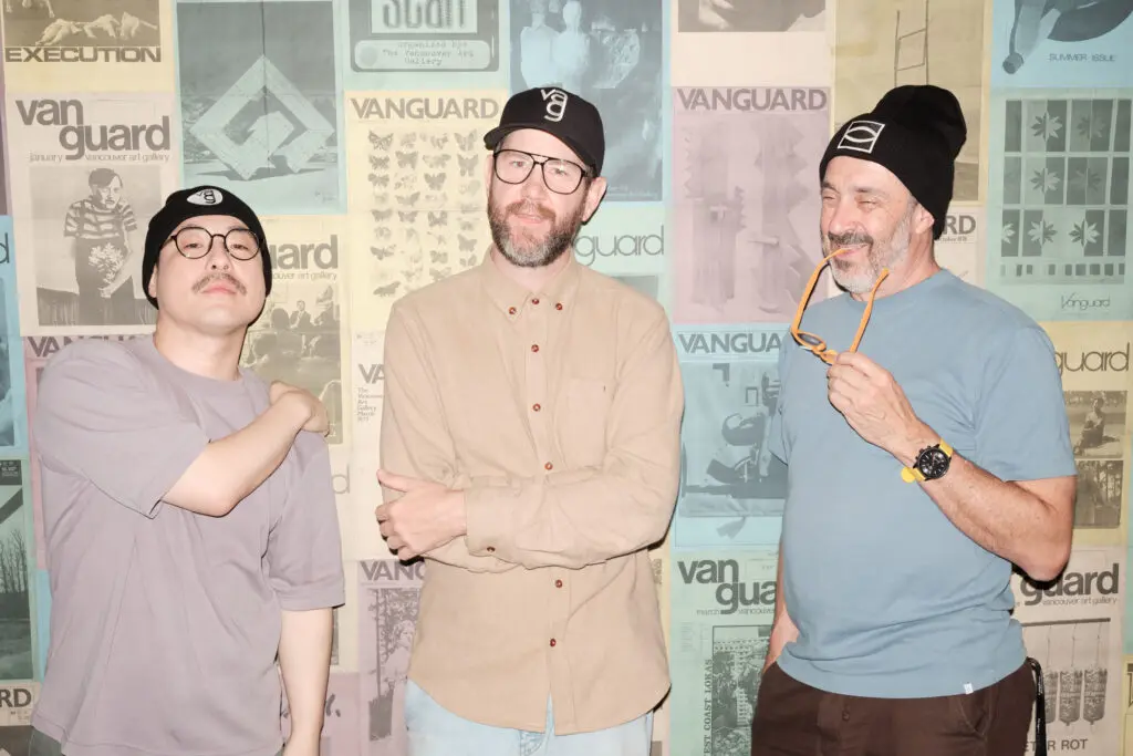 A photograph of three men wearing hats and toques. They stands in front of a wall of posters of historical magazine covers.