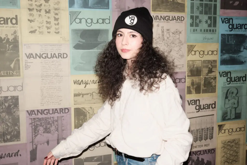 A photograph of woman with brown curly hair wearing a vag logo toque on it standing against a postered backdrop of colourful Vanguard magazine covers.