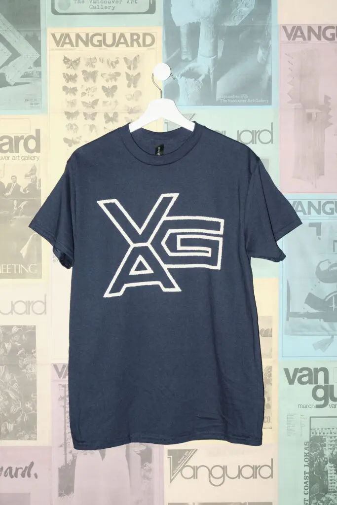 A photograph of a navy blue t-shirt with a an outline VAG logo on it hanging on a white hanger against a postered backdrop of colourful Vanguard magazine covers.