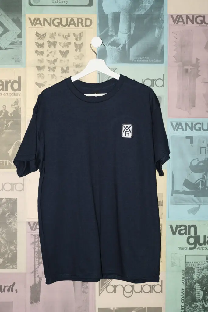 A photograph of a navy blue t-shirt with the original 1931 Gallery logo on it hanging on a white hanger against a postered backdrop of colourful Vanguard magazine covers.
