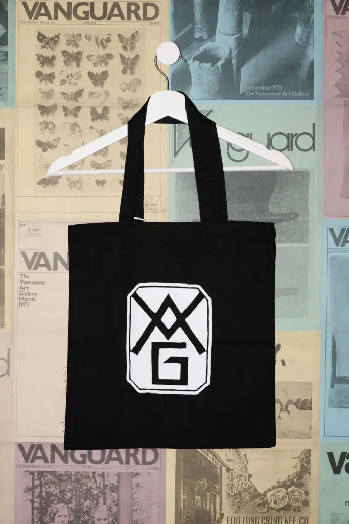 A photograph of a black tote bag featuring the Gallery's 1931 founding logo on it hanging on a white hanger against a postered backdrop of colourful Vanguard magazine covers.