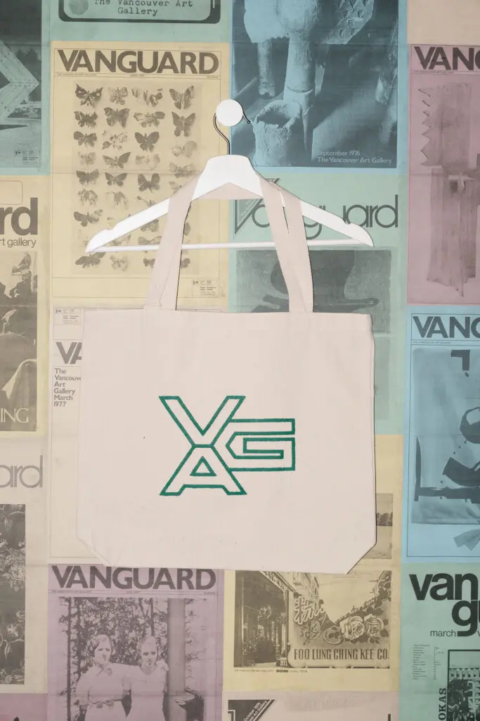 A photograph of an ecru tote bag featuring a VAG logo graphic in green on it hanging on a white hanger against a postered backdrop of colourful Vanguard magazine covers.