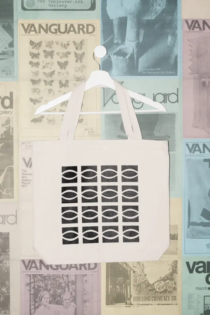 A photograph of an ecru tote bag featuring a repeated eye logo graphic on it hanging on a white hanger against a postered backdrop of colourful Vanguard magazine covers.