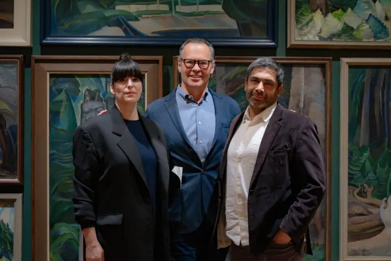 A photograph of a woman, a tall man, and another man from the torso up standing in front of paintings of trees.