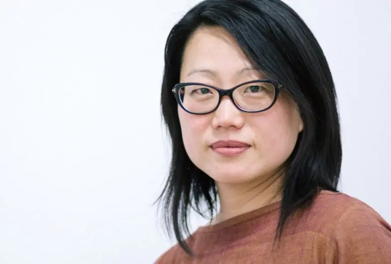 A photograph of an Asian woman wearing black glasses from the shoulders up. She has black hair and is wearing a brown shirt. The background is white.