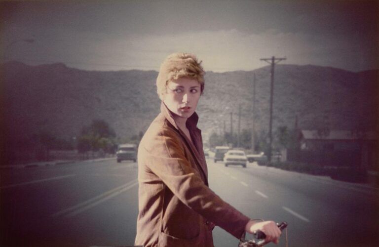A photograph by artist Cindy Sherman of herself standing in the middle of a highway, looking out at the camera in a short, curly blonde wig. The photograph looks like a film still from the 1970s.
